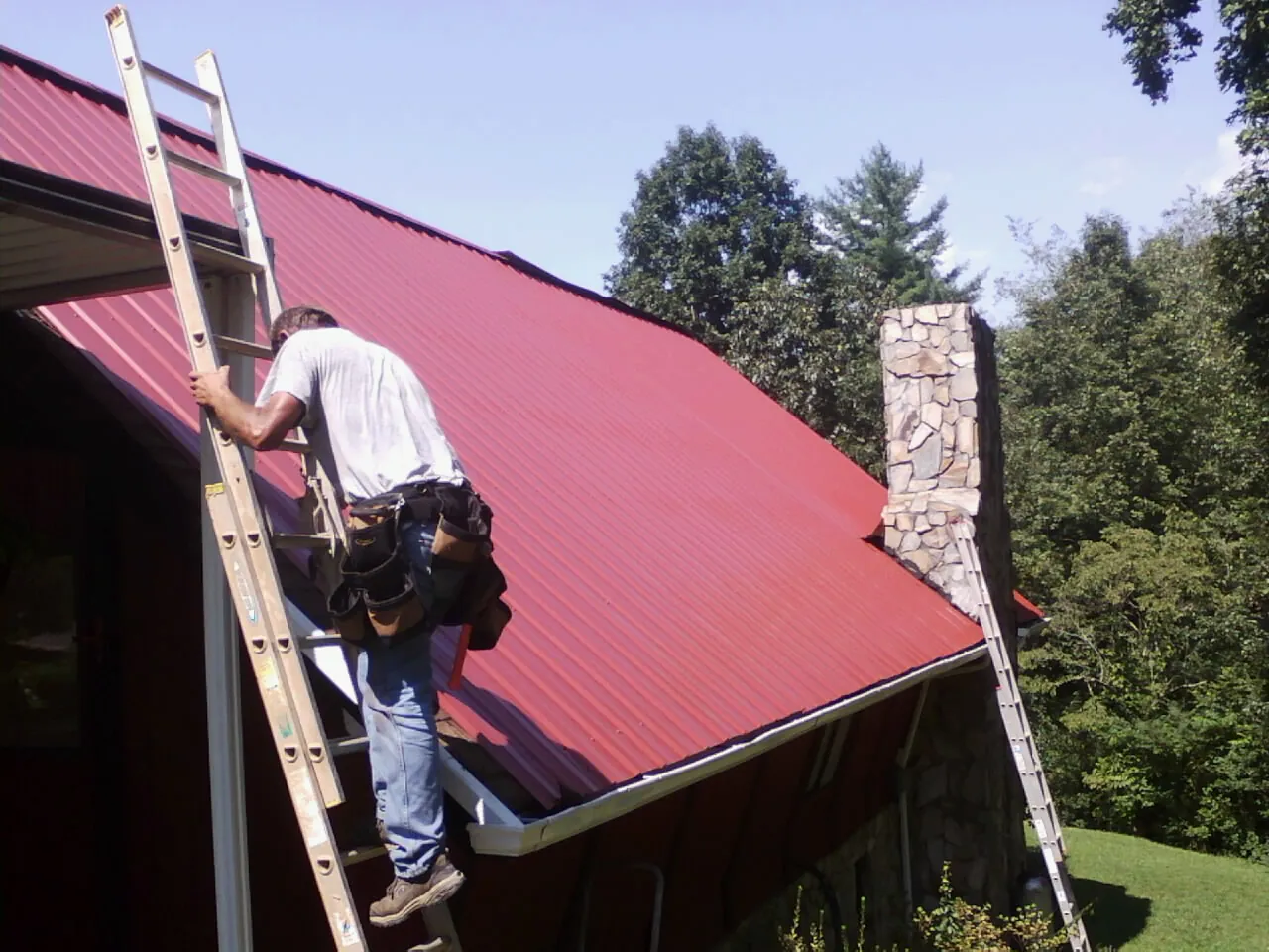 Roofer on a ladder beside a red metal roof and stone chimney — Soffit & Fascia Repair by Roofing Brothers in Clarinda